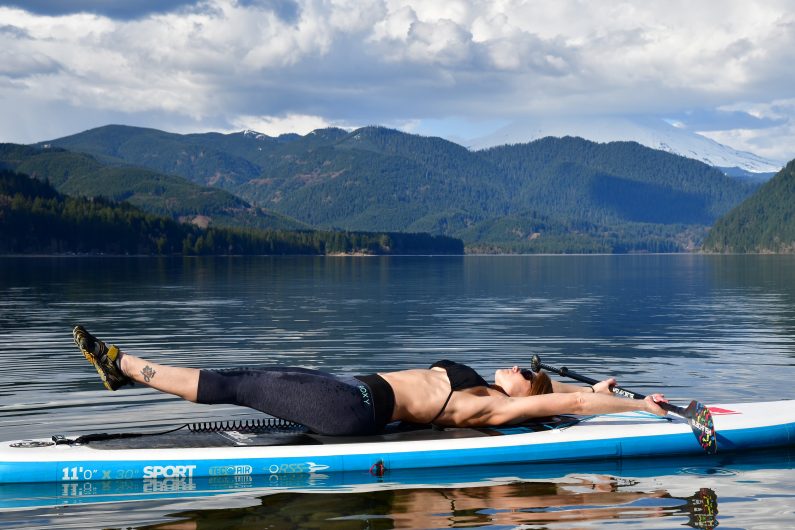 Michelle demonstrates a double leg lift, lying on her back with arms extended above her head. A serene lake below her SUP and green hills  in the distance are the backdrop.